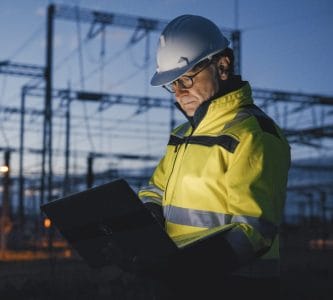 Electrical engineer in a hard hat reviewing a laptop at a power substation at dusk