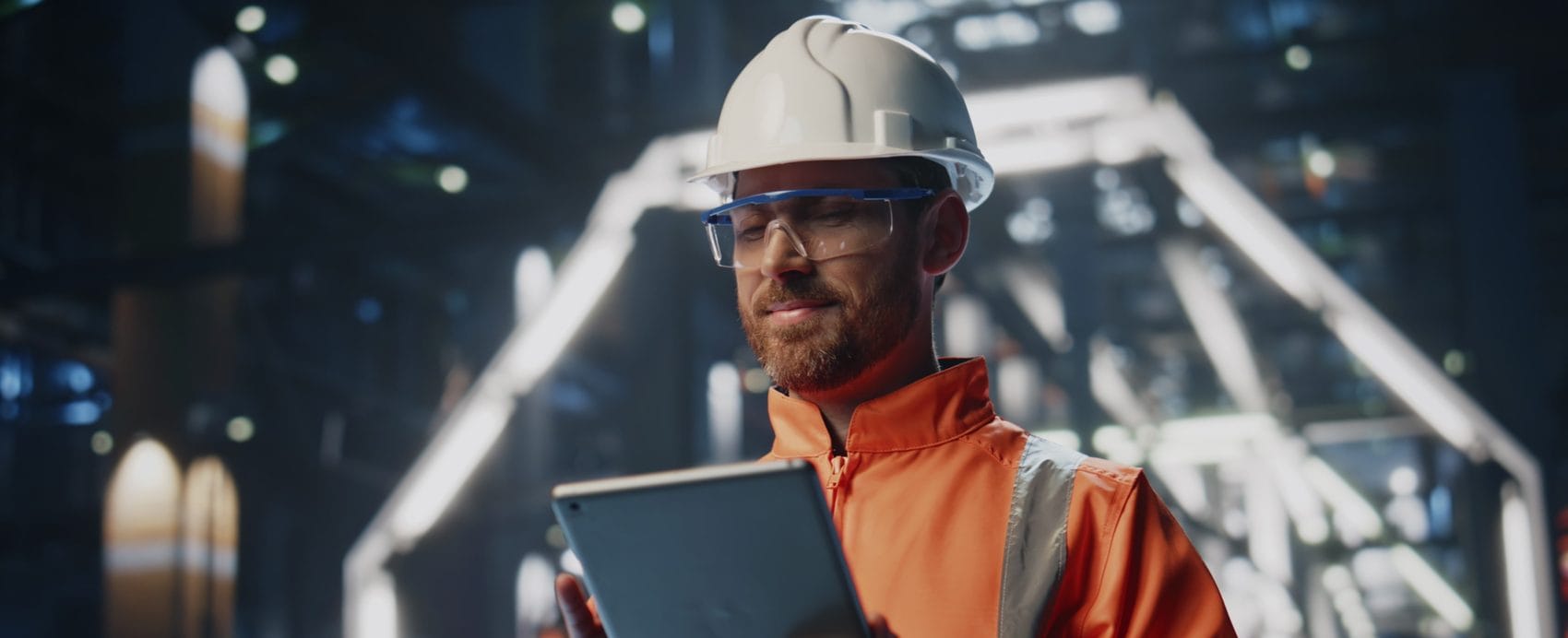 Smiling engineer in a hard hat and safety glasses checking a tablet on the production floor